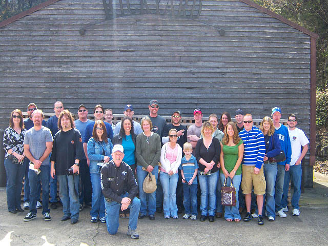 Group of people in front of a weathered wooden building, some wearing casual clothing and hats