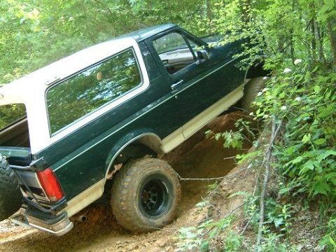 1993 Ford Bronco - A green and white Ford Bronco navigates a steep, muddy incline on a wooded trail.