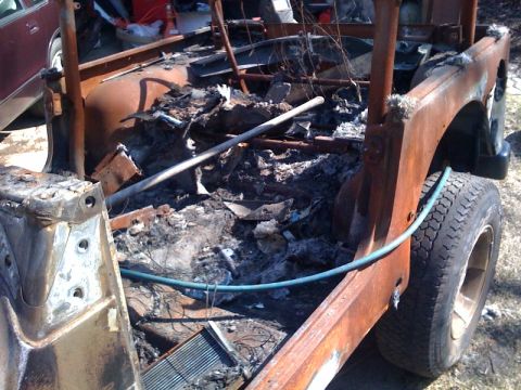 1990 Ford Bronco - rusted, burned-out bed of a vintage truck filled with charred debris, a green hose draped over the side.