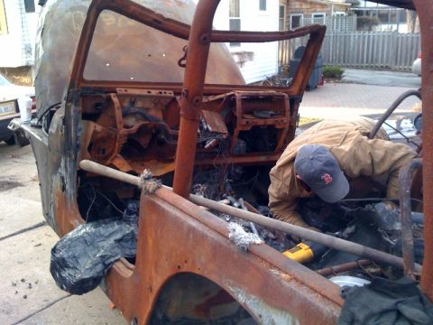 1990 Ford Bronco - person in a cap leans into the rusted, fire-damaged shell of an old vehicle being stripped in a driveway.