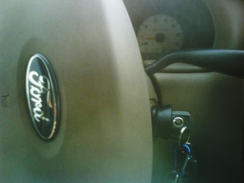 2003 Ford Taurus - Close-up of a tan steering wheel hub with a blue oval Ford emblem, ignition key in the column, and a tachometer gauge in the background.
