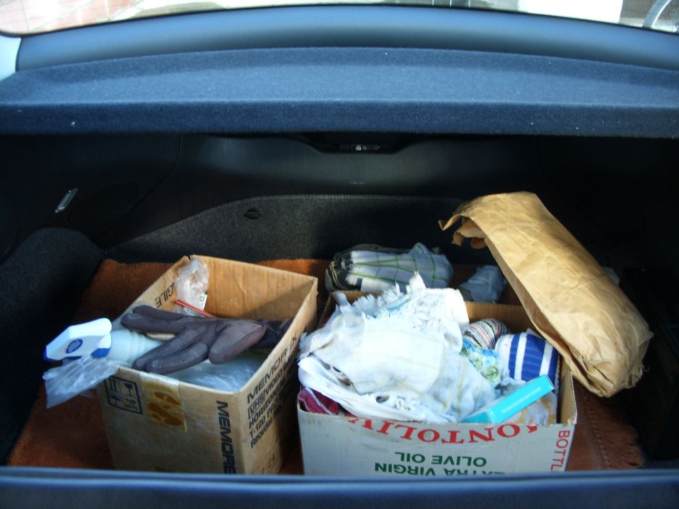 Open car trunk containing two cardboard boxes of rags, gloves, spray bottle, and a brown paper bag