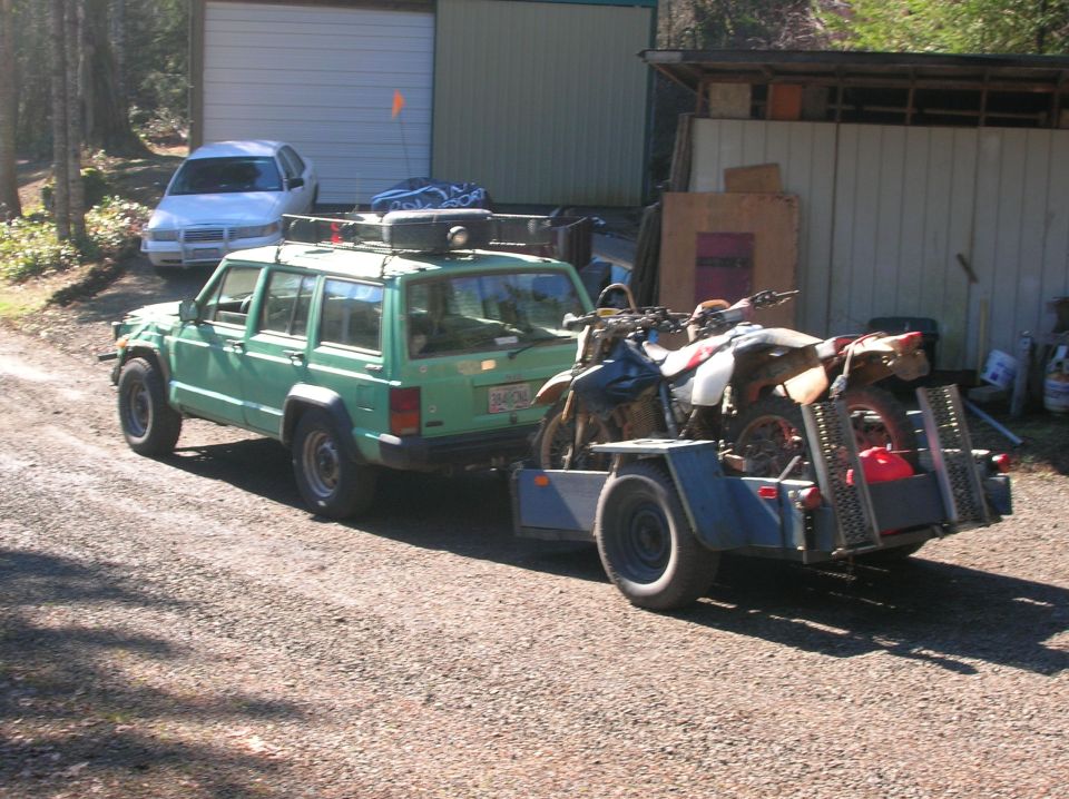 Green Jeep Cherokee with roof rack towing a trailer with two dirt bikes
