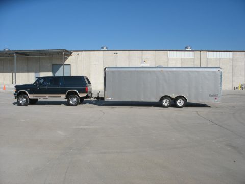 1995 Ford C-350 (Centurion Conversions) - A dark green SUV tows a large silver enclosed trailer in front of a concrete building.