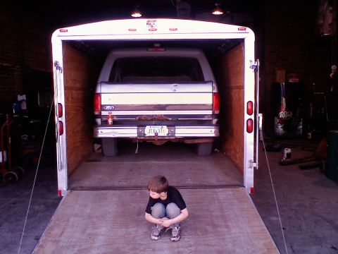 1995 Ford C-350 (Centurion Conversions) - A young boy sits on the ramp of an open trailer, looking at a Ford Bronco parked inside.