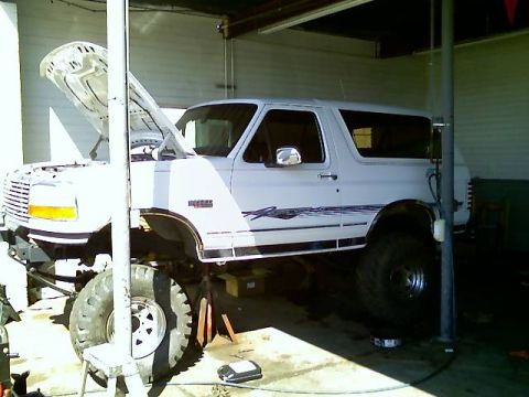 1996 Ford Bronco - A white Ford Bronco is elevated on jack stands inside a garage, with its hood open, suggesting it is undergoing maintenance or modification.