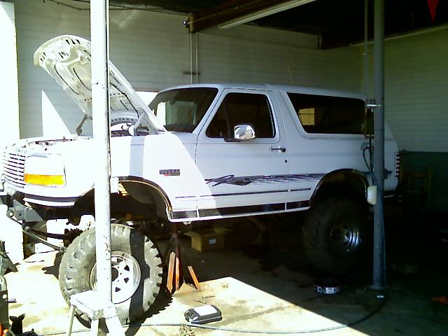 White Ford Bronco on jack stands with hood up in a garage bay, front end apart