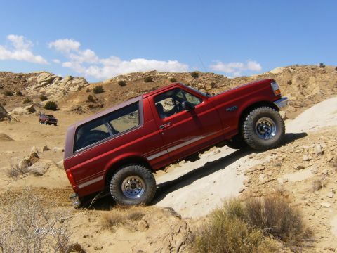 1993 Ford Bronco - A red Ford Bronco navigates a rocky, off-road terrain under a blue sky with scattered clouds.