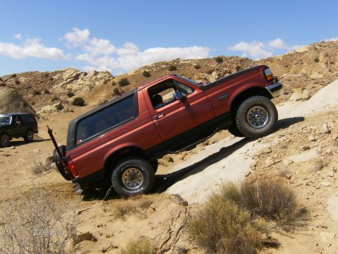 1993 Ford Bronco - A red 1993 Ford Bronco navigates a steep, rocky incline off-road, with another Bronco visible in the distance.