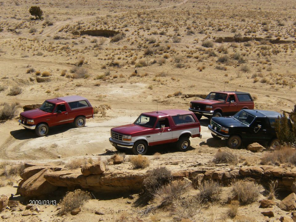 Four Ford Bronco SUVs parked in a desert landscape. The red and white Bronco in the foreground is a 1993 model