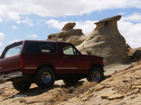 1993 Ford Bronco - A red 1993 Ford Bronco navigates a , rocky terrain under a partly cloudy sky.