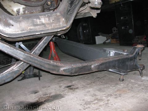 1990 Ford Bronco - Close-up of a fabricated black tubular suspension arm and skid plate mounted under a raised vehicle in a garage.