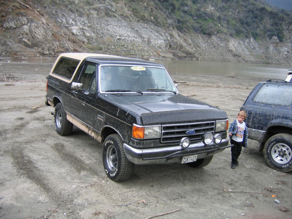 Black Ford Bronco with tan top and aftermarket lights parked on dirt near water, another SUV to the right