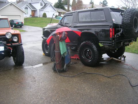 1996 Ford Bronco - Two off-road vehicles are parked on a wet driveway, with welding equipment set up near the black Bronco.