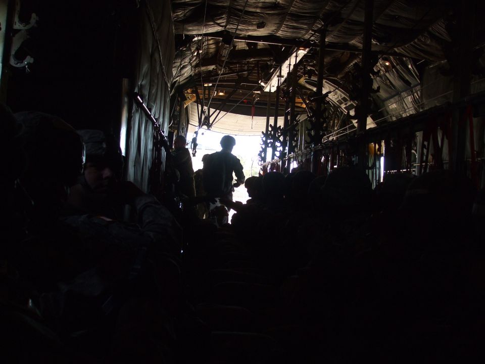 Inside a cargo plane, people are sitting and standing, silhouetted against the bright opening at the back