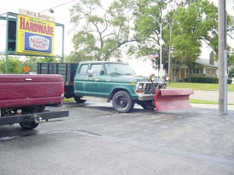 1992 Ford Bronco - A green pickup truck equipped with a snow plow is parked in front of a hardware store, with a red Ford pickup truck to its left.