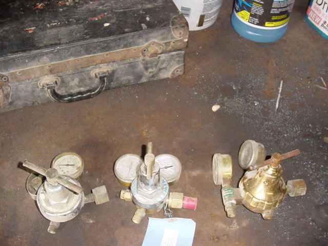 Three brass and chrome gas regulators with pressure gauges on a concrete floor by a tool case