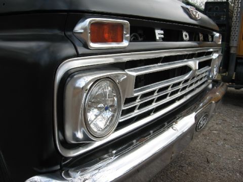 1966 Ford F-100 - Low-angle close-up of the driver-side front corner of a black Ford pickup, showing a round headlight with a blue Ford-logo lens insert, chr…