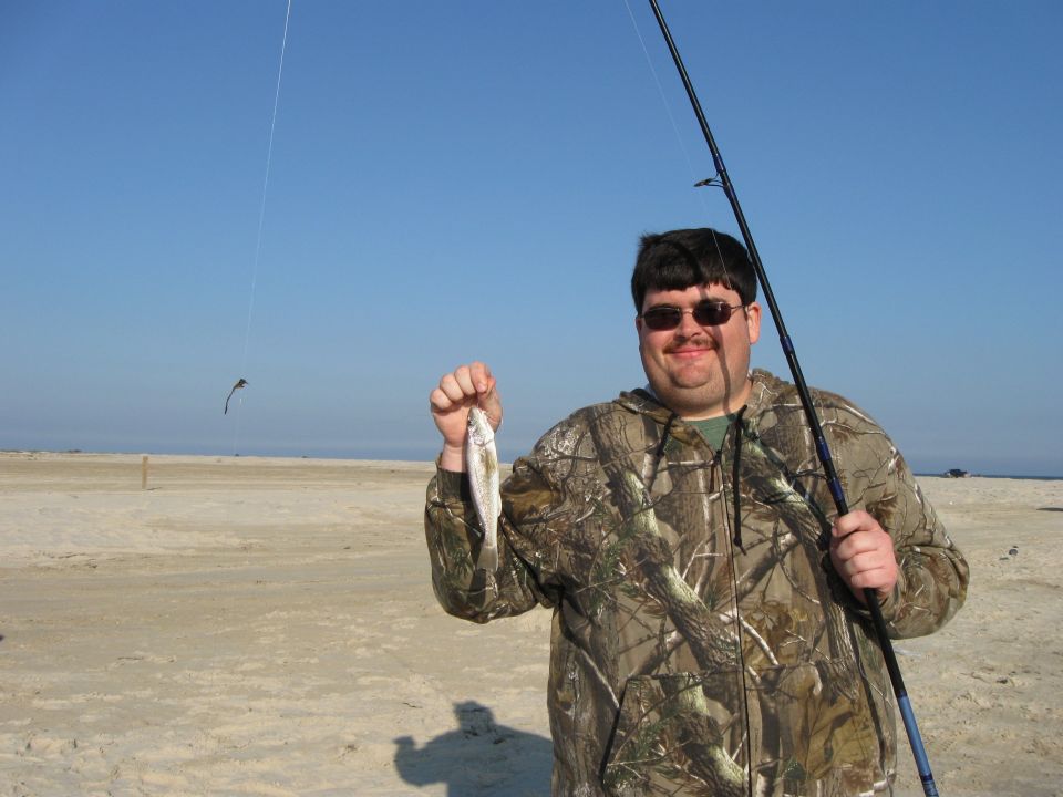 Guy on beach holding a small fish he caught, fishing rod in hand