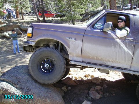 1978 Ford Bronco - A man drives a Ford Bronco over a large rock on a wooded trail, with other people and vehicles in the background.