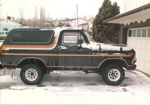 1978 Ford Bronco - A dark 1978 Ford Bronco with distinctive side stripes sits covered in snow, parked in front of a garage.