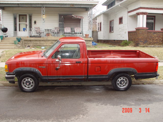 Red Ford Ranger regular-cab pickup, driver-side view, parked on street in front of white houses