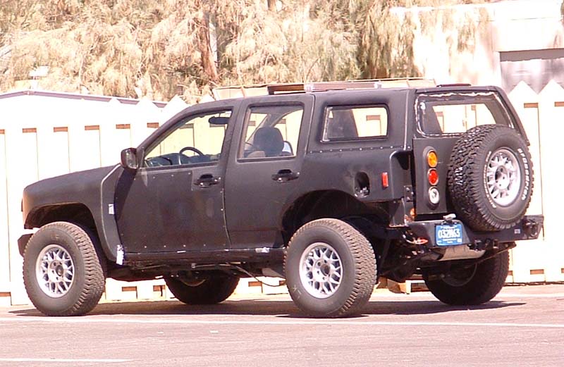 Matte black lifted SUV with rear spare tire, rear 3/4 view, parked by white fence