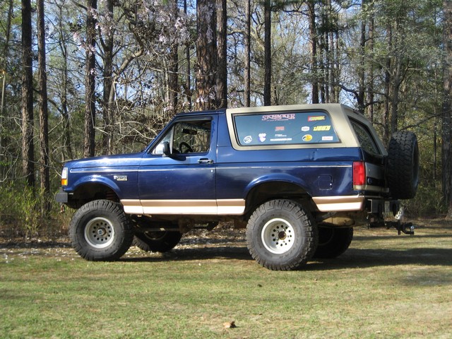 1995 Ford Bronco, lifted, two-door, blue with tan trim, parked in front of trees.
