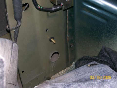 1995 Mercury Grand Marquis - A close-up view of a brass fitting installed through a hole in the olive drab painted metal interior of a vehicle. A black wire loom is vis…