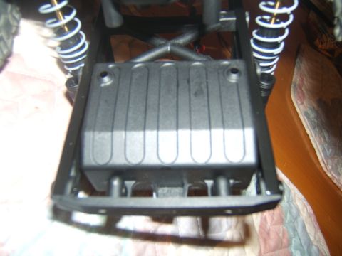 1988 Ford Bronco - The underside of an RC car chassis shows a black battery box with ribbed texture, mounted within a black frame. Visible are white and gold…