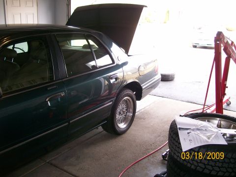 1995 Mercury Grand Marquis - A dark green sedan with its hood open is parked in a garage. A red engine hoist is visible to the right of the vehicle, along with a stack…