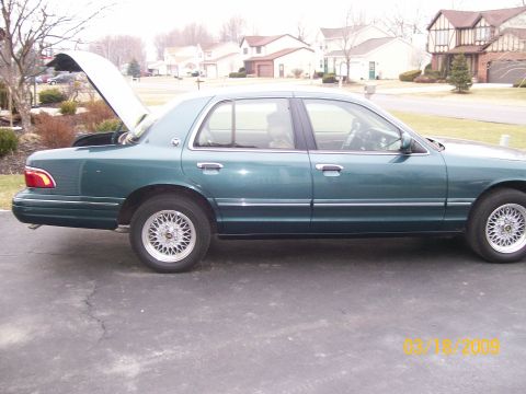 1995 Mercury Grand Marquis - The side view of a dark green sedan with the trunk open, parked on an asphalt driveway in front of houses.