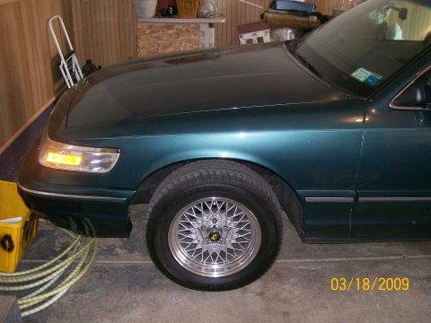 1995 Mercury Grand Marquis - The front passenger side of a dark teal sedan is shown, featuring a chrome grille, amber turn signal, and a wire-spoke wheel. The vehicle i…