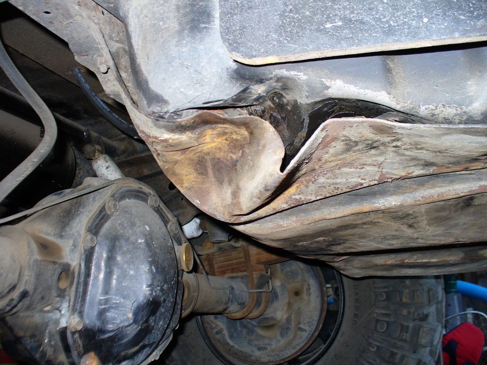 Underside of a vehicle showing a damaged and rusted fuel tank area, differential, and tire.