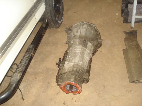 1991 Ford Bronco - A transmission is lying on the ground next to the running board and tire of a white vehicle. A metal skid plate is also visible on the grou…