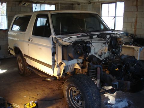 1991 Ford Bronco - A white Ford Bronco is parked inside a garage with its front end disassembled. The vehicle is missing its hood and fenders, revealing the e…