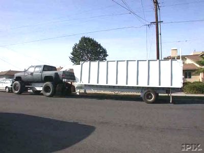 1978 Ford Bronco - A lifted pickup truck tows a large white trailer down a residential street.