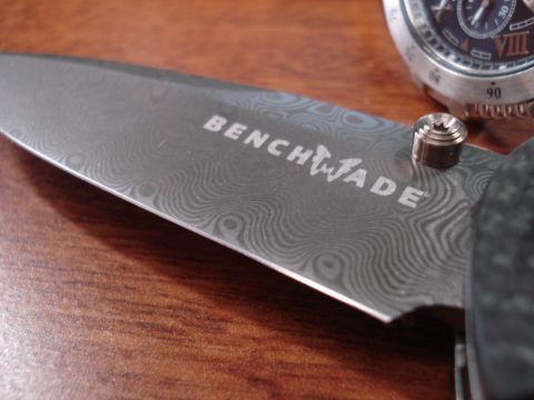 2000 Ford Crown Victoria - A close-up shot of a knife with the "BENCHMADE" logo etched on the blade. The blade has a Damascus pattern and is resting on a wooden surfa…
