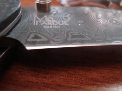 2000 Ford Crown Victoria - A close-up shot of a knife blade with a Damascus pattern and engraved text that reads "MEL TARDUE DESIGN". The blade is resting on a wooden…