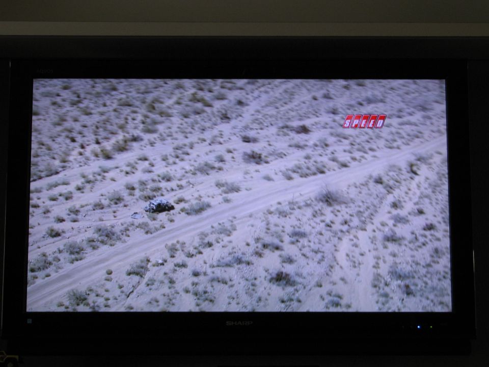 Aerial view of a desert scene with a dirt road and a small vehicle, featuring a SPEED logo.