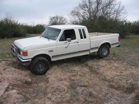 1990 Ford F-150 - A white 1990 Ford F-150 pickup truck is shown from a 3/4 front angle, partially stuck in a muddy ditch. The truck is in an outdoor, grassy…