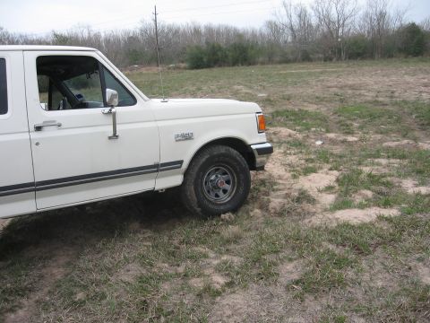 1990 Ford F-150 - The right side of a white 1990 Ford F-150 pickup truck is parked on a dirt and grass lot. The front wheel and part of the door are visible.