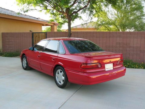 1995 Ford Taurus - A red 1995 Ford Taurus sedan is parked in front of a brick wall and a house.