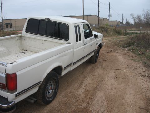1990 Ford F-150 - The rear passenger side of a white Ford pickup truck is shown parked on a dirt road. The truck bed is empty, and the vehicle is facing away…