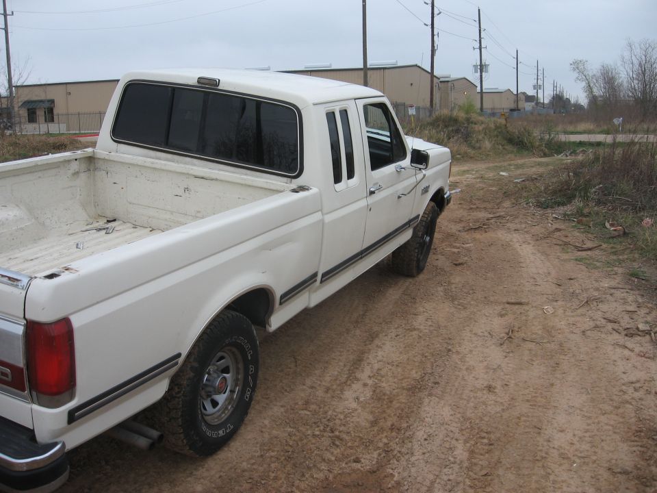White Ford pickup truck, rear passenger side view, parked on dirt road.