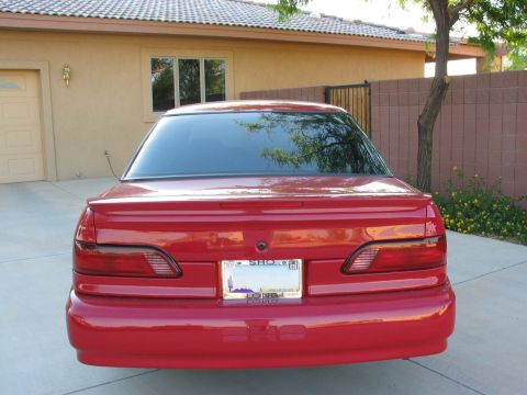 1995 Ford Taurus - The rear of a red sedan is parked in front of a house, showing its taillights and license plate.