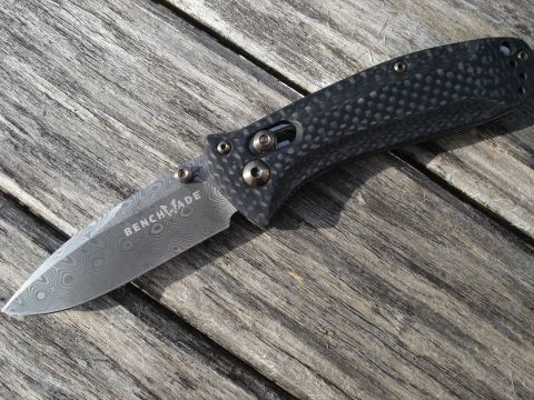 2000 Ford Crown Victoria - A close-up, angled shot of a folding knife with a Damascus steel blade and a carbon fiber handle resting on a weathered wooden surface.