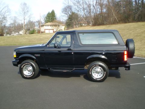 1996 Ford Bronco - The side view of a black 1996 Ford Bronco is shown parked on an asphalt surface with a grassy hill and houses in the background. The vehicl…