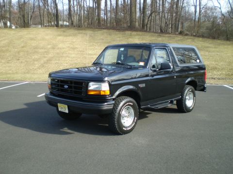 1996 Ford Bronco - A black 1996 Ford Bronco is parked on an asphalt lot with a grassy hill and trees in the background. The vehicle is shown from a 3/4 front…