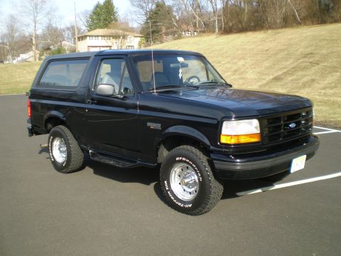 1996 Ford Bronco - A black 1996 Ford Bronco is parked on an asphalt surface with a grassy hill and houses in the background. The SUV is shown from a 3/4 front…